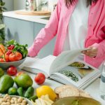 A person browsing a cookbook surrounded by fresh vegetables in a bright kitchen setting.
