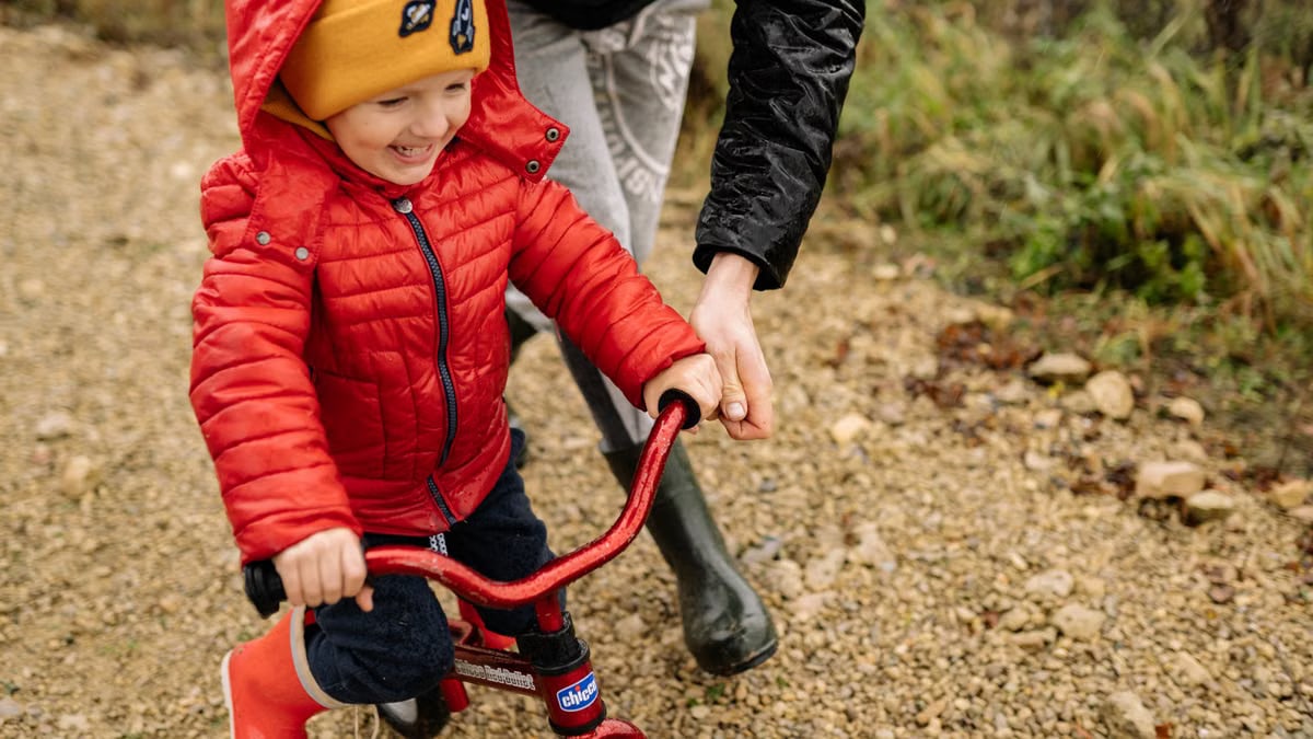Joyful child riding a bike with parent on a gravel path, enjoying family time outdoors.